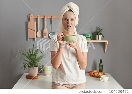 Photo of scared Caucasian woman with towel on head standing speechless indoor with beauty facial mask to reduce fine lines eats her breakfast, awakes early in morning, posing in kitchen. 104330421