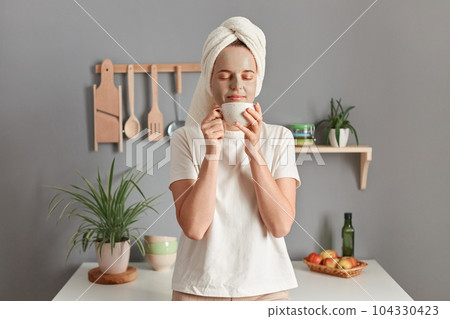 Indoor shot of calm relaxed young adult woman wrapped white towel and dressed in T-shirt standing near table on kitchen at home, smelling coffee, enjoying aromat, keeps eyes closed. Indoor shot of calm relaxed young adult woman wrapped white towel and dressed in T-shirt standing near table on kitchen at home, smelling coffee, enjoying aromat, keeps eyes closed. 104330423