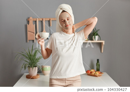 Indoor shot of young adult attractive woman with cleansing mask on her face standing in the kitchen at home, drinking her morning coffee, wakes up early. 104330427