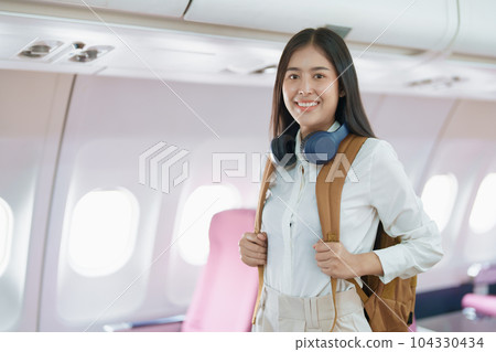 Young asian attractive woman travel by airplane, Passenger wearing headphone putting hand baggage in lockers above seats of plane. 104330434