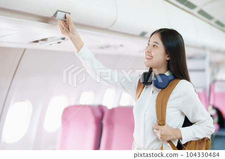 Young asian attractive woman travel by airplane, Passenger wearing headphone putting hand baggage in lockers above seats of plane. 104330484