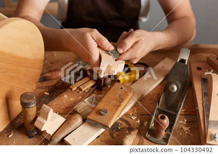 Closeup of man carpenter wearing white t-shirt, black cap and brown apron working, uses chisel for cutting shapes and curves in wood sits at table surrounded by different instruments. 104330492