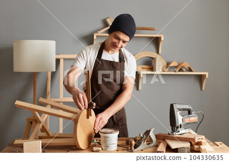 Indoor shot of caucasian young adult man carpenter wearing white t-shirt, black cap and brown apron working, making wooden chair, enjoying his hobby and job. Indoor shot of caucasian young adult man carpenter wearing white t-shirt, black cap and brown apron working, making wooden chair, enjoying his hobby and job. 104330635