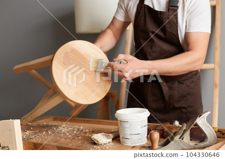 Closeup portrait of faceless anonymous carpenter wearing brown apron applies paint using paintbrush, decorating wooden chair in carpentry workshop, working. 104330646