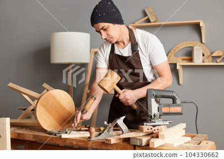 Portrait of master wearing brown apron and black cap using mallet and chisel to smooth rectangular block of wood while it stands on top of table, having concentrated facial expression. Portrait of master wearing brown apron and black cap using mallet and chisel to smooth rectangular block of wood while it stands on top of table, having concentrated facial expression. 104330662