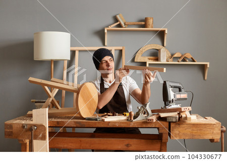 Portrait of hard working concentrated woodworker, young male carpenter sitting at the wooden table with diverse working tools, making wooden furniture, makes chair. 104330677