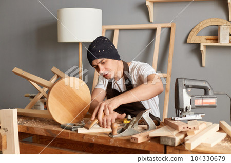 Indoor shot of concentrated young adult man carpenter wearing black cap and brown apron sanding wooden wooden block in his workshop while sitting at table in his workshop. 104330679
