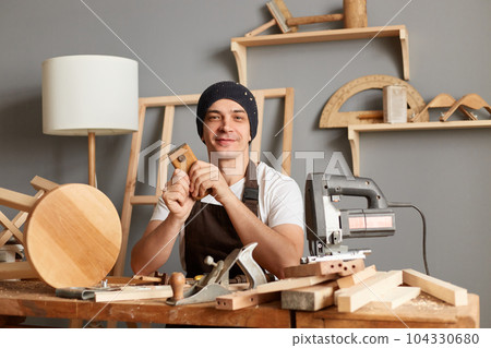 Portrait of positive optimistic young adult man carpenter sanding wooden wooden block in his workshop, sitting at table looking at camera, enjoying his work in joinery. Portrait of positive optimistic young adult man carpenter sanding wooden wooden block in his workshop, sitting at table looking at camera, enjoying his work in joinery. 104330680