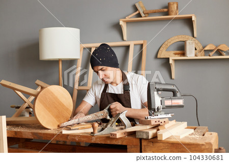 Indoor shot of carpenter man using saw hand cutting wooden plank in workshop, making wooden products, sitting at table in joinery, wearing cap and apron. 104330681