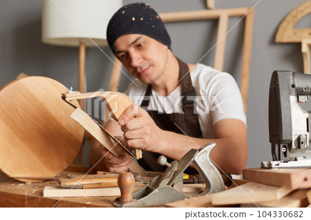 Image of calm attractive young adult man carpenter wearing white t-shirt, black cap and brown apron working in joinery, making wooden chair, sanding wood block. Image of calm attractive young adult man carpenter wearing white t-shirt, black cap and brown apron working in joinery, making wooden chair, sanding wood block. 104330682