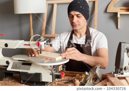 Portrait of attractive caucasian man carpenter wearing brown apron and black cap, using electric jigsaw, working in his joinery, enjoying process of making wooden products. Portrait of attractive caucasian man carpenter wearing brown apron and black cap, using electric jigsaw, working in his joinery, enjoying process of making wooden products. 104330726