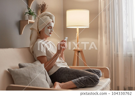 Attractive young Caucasian woman wrapped in towel and facial clay mask on her face, using her mobile phone while relaxing on sofa in her living room, doing cosmetology procedures Attractive young Caucasian woman wrapped in towel and facial clay mask on her face, using her mobile phone while relaxing on sofa in her living room, doing cosmetology procedures 104330762