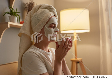 Side view portrait of happy delighted woman with cosmetology mask on face sitting on cough, holding cup of coffee and enjoying aromat, relaxing at home. Side view portrait of happy delighted woman with cosmetology mask on face sitting on cough, holding cup of coffee and enjoying aromat, relaxing at home. 104330770
