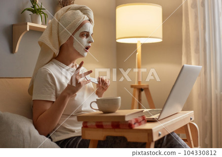 Indoor shot of shocked scared woman wearing white T-shirt and towel sitting on sofa at home, doing skin care procedures and manicure, watching horror film or shock content in internet. 104330812