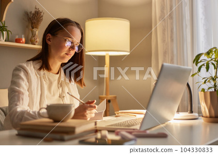 Indoor shot of focused woman wearing white shirt sitting on table and working on laptop near window, having concentrated facial expression, studying hard or works with a new project. 104330833