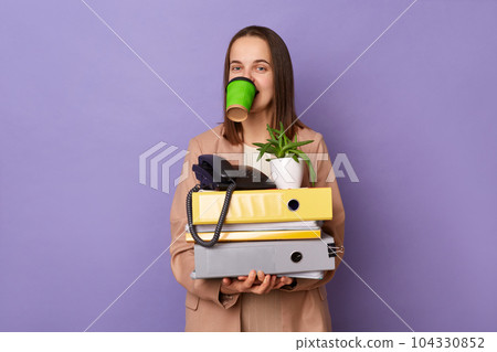 Portrait of funny woman wearing beige jacket holding lot of documents folders isolated over purple background, holds takeaway coffee in her mouth, having lots work. 104330852