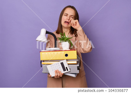 Image of crying unhappy woman wearing beige jacket holding lot of documents folders isolated over purple background, being sad, loosing her job, standing with her stuff in hands. Image of crying unhappy woman wearing beige jacket holding lot of documents folders isolated over purple background, being sad, loosing her job, standing with her stuff in hands. 104330877