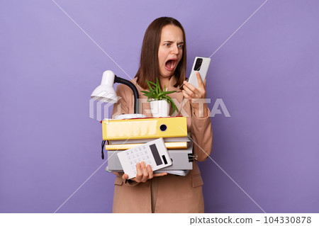Portrait of scared shocked woman wearing beige jacket holding lot of documents folders, holding smart phone, looking at display and screaming, having bad news, posing isolated over purple background. Portrait of scared shocked woman wearing beige jacket holding lot of documents folders, holding smart phone, looking at display and screaming, having bad news, posing isolated over purple background. 104330878