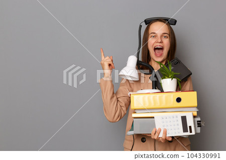 Portrait of joyful cheerful woman wearing beige jacket, holding paper folders posing isolated over gray background, pointing aside at copy space for promotional text. 104330991