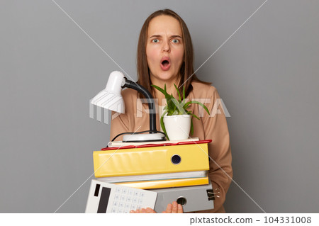 Indoor shot of shocked astonished surprised young pretty woman wearing beige jacket holding documents and her office stuff posing isolated over gray background, looking at camera with big eyes. Indoor shot of shocked astonished surprised young pretty woman wearing beige jacket holding documents and her office stuff posing isolated over gray background, looking at camera with big eyes. 104331008