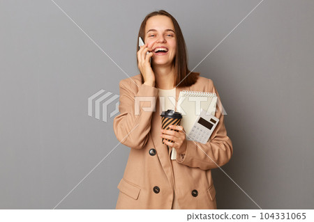 Indoor shot of amazed excited woman with brown hair wearing beige jacket, talking on cell phone and laughing, holding her work stuff, standing isolated over gray background. 104331065