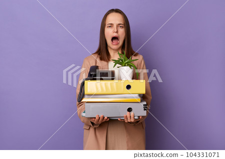 Portrait of despair young adult woman wearing beige jacket holding lot of documents folders, screaming, expressing negative emotions, posing isolated over lilac background. 104331071