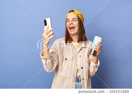 Photo of joyful cheerful brown haired teenager girl wearing baseball cap and jacket posing isolated over blue background, using mobile phone while drinking hot coffee, laughing, livestreaming. 104331088