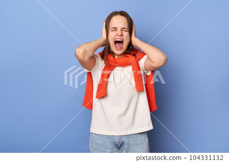 Portrait of annoyed frustrated woman wearing casual style clothing standing isolated over blue background, screaming with despair expression, covering her ears. Portrait of annoyed frustrated woman wearing casual style clothing standing isolated over blue background, screaming with despair expression, covering her ears. 104331132
