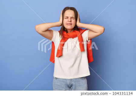 Portrait of despair dark haired woman wearing white T-shirt and orange sweater tied over shoulders, covering ears with hands, avoids noise, posing isolated on blue background. Portrait of despair dark haired woman wearing white T-shirt and orange sweater tied over shoulders, covering ears with hands, avoids noise, posing isolated on blue background. 104331134
