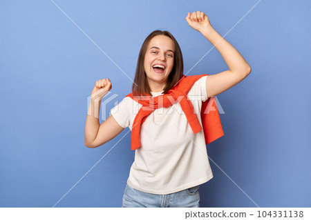Portrait of extremely happy smiling woman wearing casual style clothing, posing with raised arms, expressing positive emotions, celebrating, standing isolated over blue background. 104331138