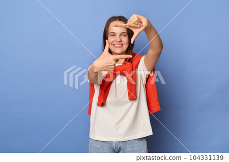 Portrait of cheerful optimistic woman photographer wearing casual style clothing standing isolated over blue background, looking at camera through frame from fingers, focus. 104331139