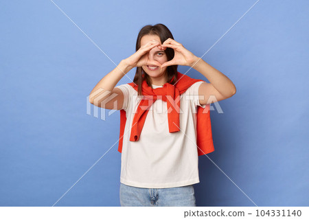 Photo of romantic flirting friendly young adult woman dressed white T-shirt and orange jumper tied over shoulders, showing heart shape with hands, standing against blue wall. 104331140