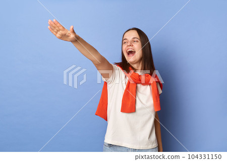 Photo of excited happy smile young adult woman dressed white T-shirt and orange jumper tied over shoulders standing against blue wall, standing with raised arms, screaming. Photo of excited happy smile young adult woman dressed white T-shirt and orange jumper tied over shoulders standing against blue wall, standing with raised arms, screaming. 104331150