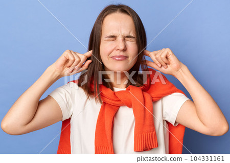 Frustrated annoyed woman in T-shirt and sweater tied over shoulders, covering ears with hands, feeling irritated with loud annoying noise, can't concentrate on her work, isolated on blue background. Frustrated annoyed woman in T-shirt and sweater tied over shoulders, covering ears with hands, feeling irritated with loud annoying noise, can't concentrate on her work, isolated on blue background. 104331161