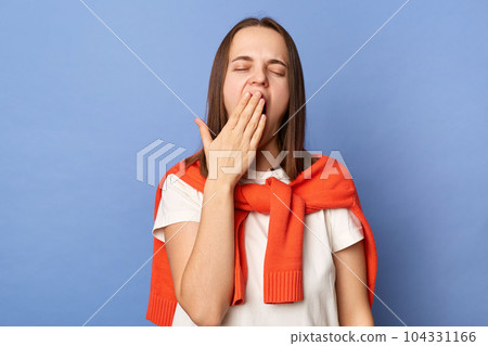 Sleepy woman wearing white T-shirt and sweater tied over shoulders, yawns as being tired and needs good rest after hard working day, gets up early in morning, standing isolated on blue background. 104331166