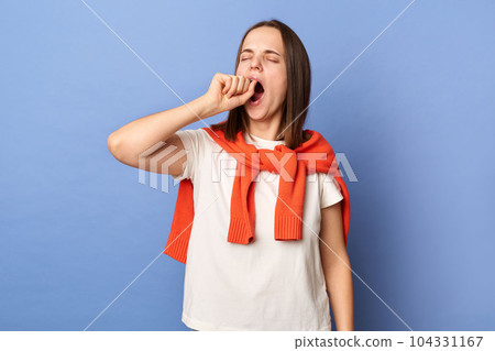 Indoor shot of sleepless tired exhausted woman dressed white T-shirt and tied sweater over shoulders standing isolated on blue background, yawning, covering mouth with fist, needs to have nap. 104331167
