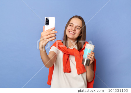Image of smiling delighted Caucasian woman blogger wearing white T-shirt and orange sweater tied over shoulders standing isolated on blue background, taking selfie, making content for her blog. Image of smiling delighted Caucasian woman blogger wearing white T-shirt and orange sweater tied over shoulders standing isolated on blue background, taking selfie, making content for her blog. 104331193