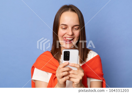 Indoor shot of positive woman wearing white T-shirt and orange sweater tied over shoulders standing isolated on blue background, using cell phone, smiling happily, sticking tongue. 104331203
