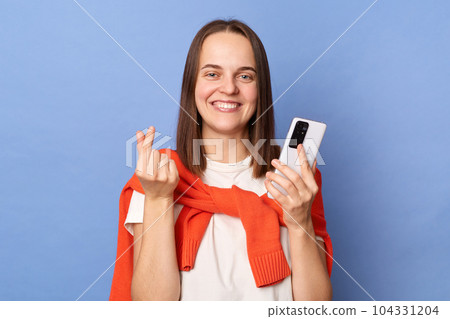 Image of charming smiling Caucasian woman wearing white T-shirt and orange sweater tied over shoulders standing isolated on blue background, using mobile phone, showing love gesture with fingers. Image of charming smiling Caucasian woman wearing white T-shirt and orange sweater tied over shoulders standing isolated on blue background, using mobile phone, showing love gesture with fingers. 104331204