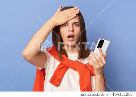 Photo of shocked young adult woman wearing white T-shirt and orange sweater tied over shoulders standing isolated on blue background, holding cell phone, touches her forehead, having problems. Photo of shocked young adult woman wearing white T-shirt and orange sweater tied over shoulders standing isolated on blue background, holding cell phone, touches her forehead, having problems. 104331205