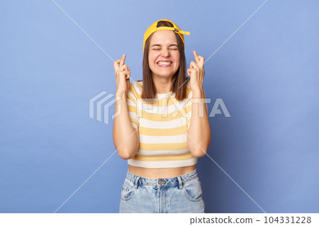 Portrait of smiling delighted caucasian teen girl wearing striped T-shirt and baseball cap standing isolated over blue background, crossing fingers, keeps eyes closed, smiling toothily. Portrait of smiling delighted caucasian teen girl wearing striped T-shirt and baseball cap standing isolated over blue background, crossing fingers, keeps eyes closed, smiling toothily. 104331228