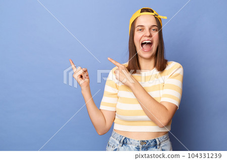 Photo of extremely happy amazed teenager girl wearing striped T-shirt and baseball cap, pointing at empty wall, showing copy space, advertisement area, posing isolated over blue background, Photo of extremely happy amazed teenager girl wearing striped T-shirt and baseball cap, pointing at empty wall, showing copy space, advertisement area, posing isolated over blue background, 104331239