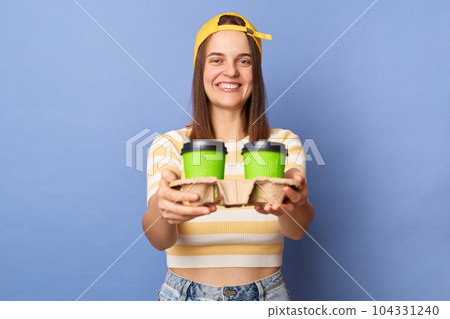 Portrait of positive teenager girl wearing striped T-shirt and baseball cap posing isolated over blue background, female working as courier, delivering takeaway coffee. Portrait of positive teenager girl wearing striped T-shirt and baseball cap posing isolated over blue background, female working as courier, delivering takeaway coffee. 104331240
