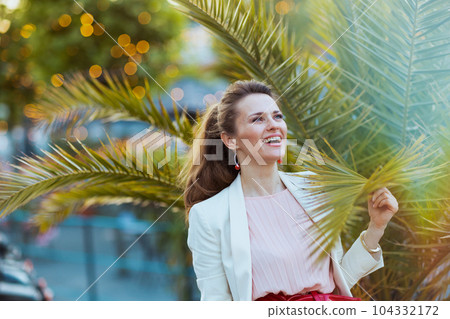 smiling elegant female in pink dress and white jacket in city 104332172