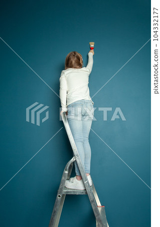 Child girl in jeans painting blue wall, doing renovation with brush and standing on the ladder Child girl in jeans painting blue wall, doing renovation with brush and standing on the ladder 104332177