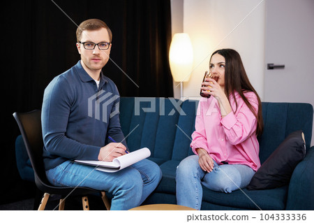 A young psychologist conducts a psychotherapy session for a woman who has psychological problems. A woman sits on a sofa and holds a glass of water in her hands 104333336