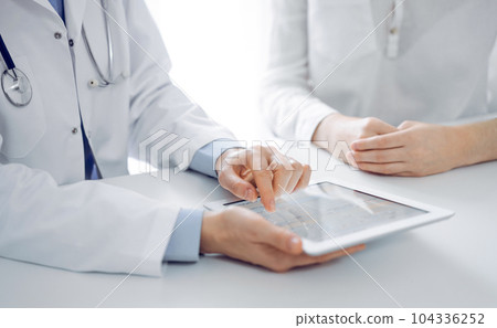 Doctor and patient sitting near each other at the desk in clinic. The focus is on female physician's hands pointing into tablet computer touchpad, close up. Medicine concept 104336252