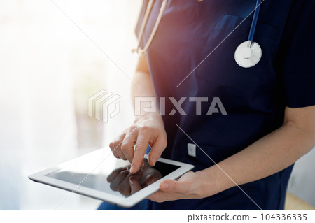 Doctor woman using tablet computer while standing near panorama window in clinic, close up. Physician or surgeon at work. Medicine concept 104336335