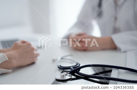 Doctor and patient sitting and discussing something at the desk in clinic. The focus is on the stethoscope lying on the table, close up. Medicine and healthcare concept 104336388