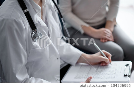 Doctor and patient sitting at sofa in clinic office. The focus is on female physician's hands filling up the medication history record form, close up. Medicine concept Doctor and patient sitting at sofa in clinic office. The focus is on female physician's hands filling up the medication history record form, close up. Medicine concept 104336995